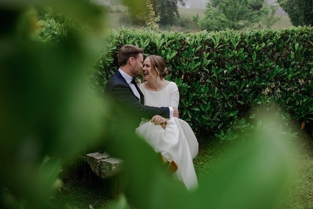 A couple sitting on a bench amongst manicured shrubs of Mali Brae Farm. They are looking at each other and laughing.