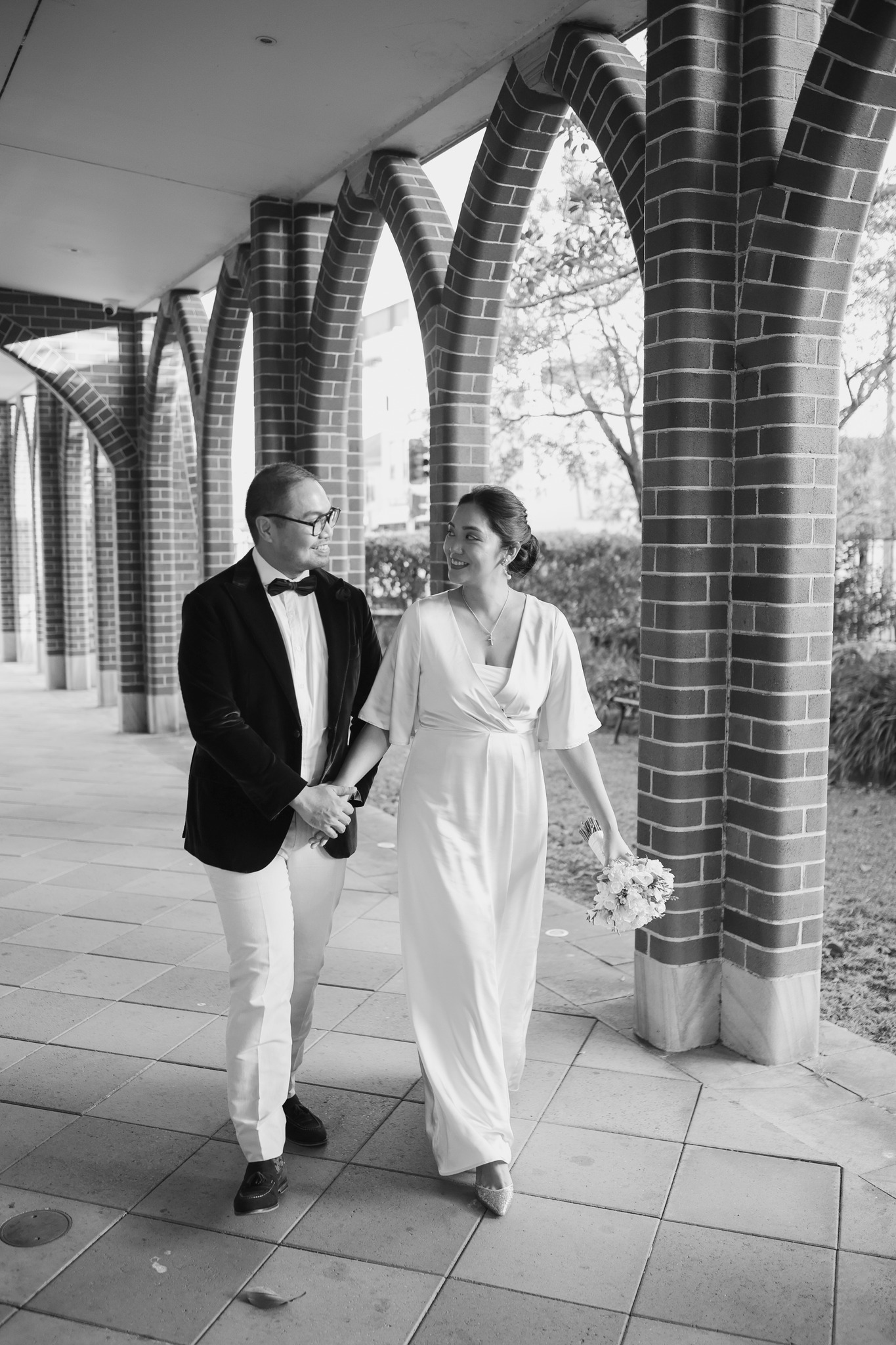 A just-married couple holding hands, walking against brick arches.